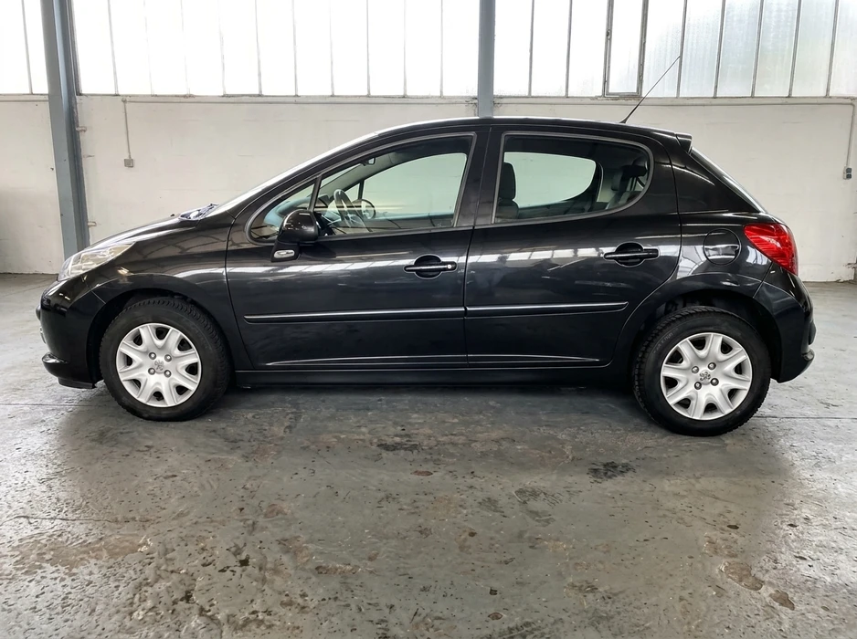 Véhicule Peugeot 207 cinq portes, coloris noir brillant, photographié de profil dans un espace intérieur industriel