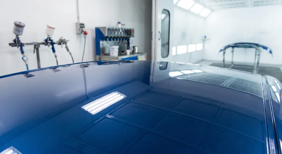 Gros plan ou vue détaillée d'une carrosserie automobile partiellement peinte ou fraîchement repeinte, montrant la qualité brillante et l'homogénéité de la peinture à teinte parfaitement respectée. La surface doit refléter la lumière uniformément, illustrant la précision colorimétrique et la finition professionnelle. Contexte d'atelier automobile intérieur visible en arrière-plan avec équipements de peinture professionnels.