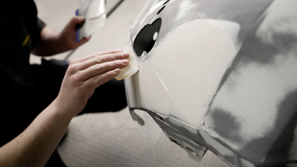 Un technicien automobile portant des gants noirs utilise une polisseuse orbitale sur le phare avant d'une voiture noire dans un atelier de carrosserie